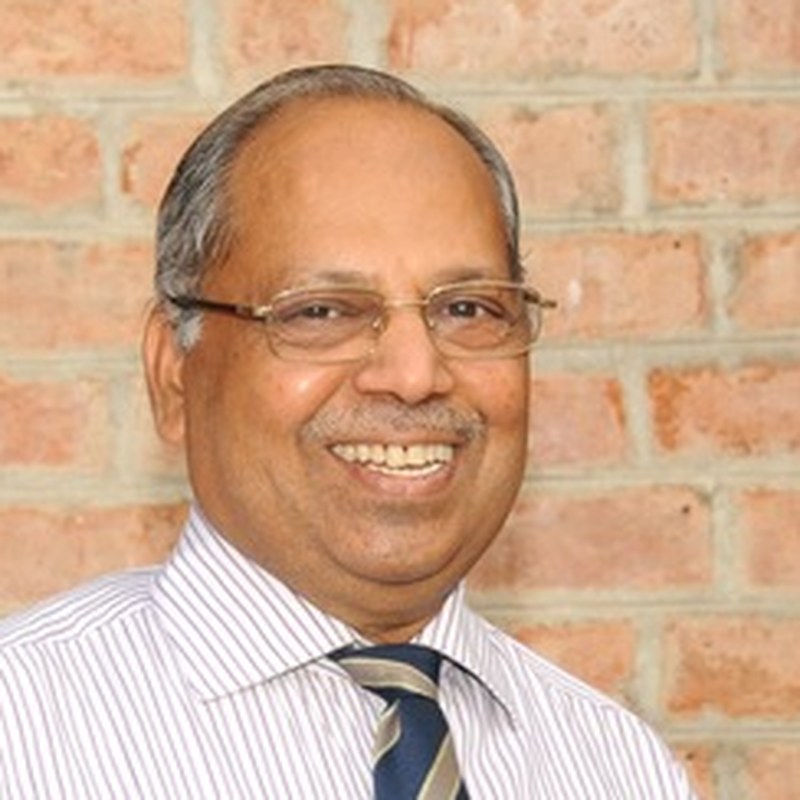 Dr. T.V. Rao, Coacharya board member, smiling in a striped shirt and tie against a brick wall