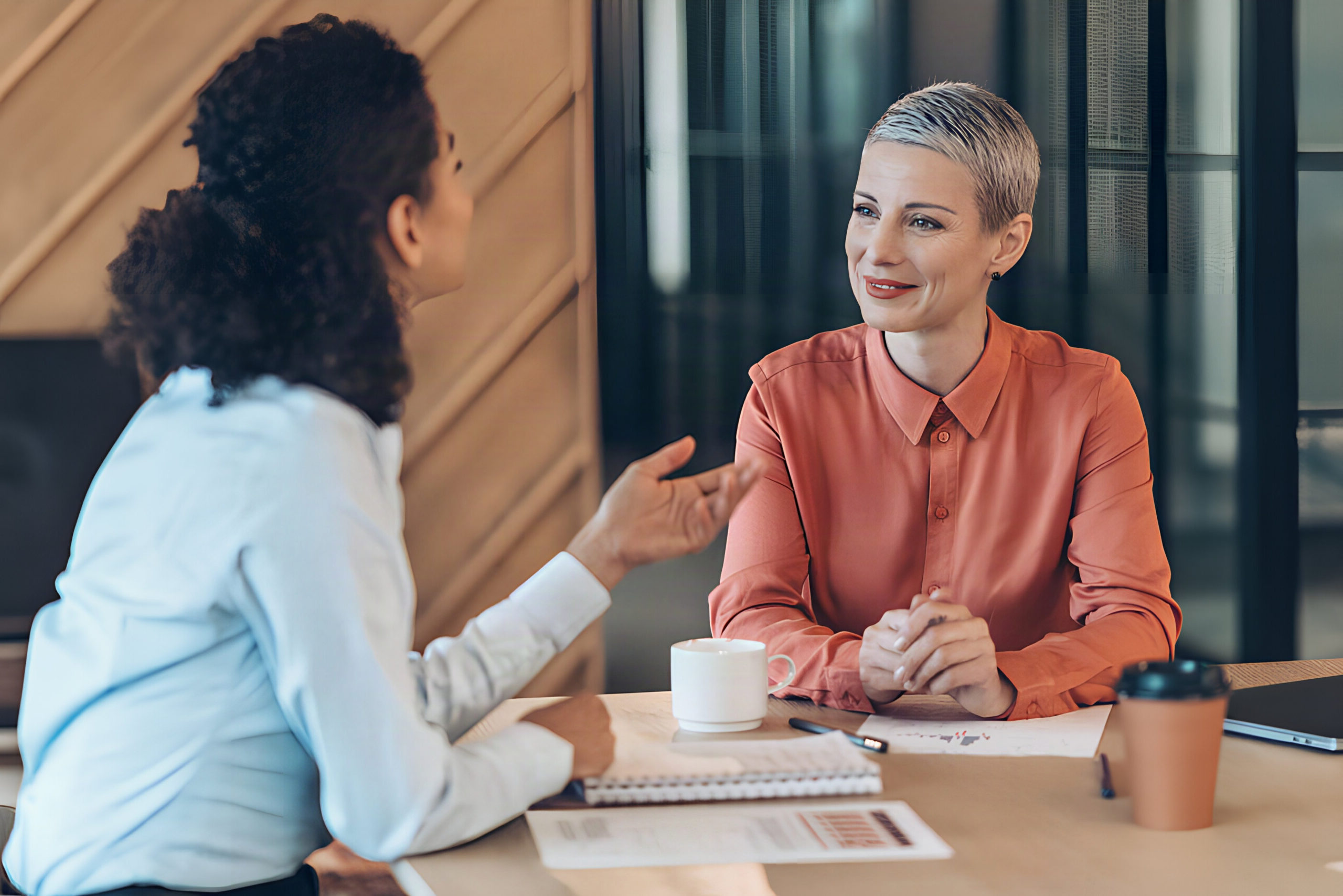 Manager in conversation with a team member across a meeting room table, listening and gesturing as she speaks.