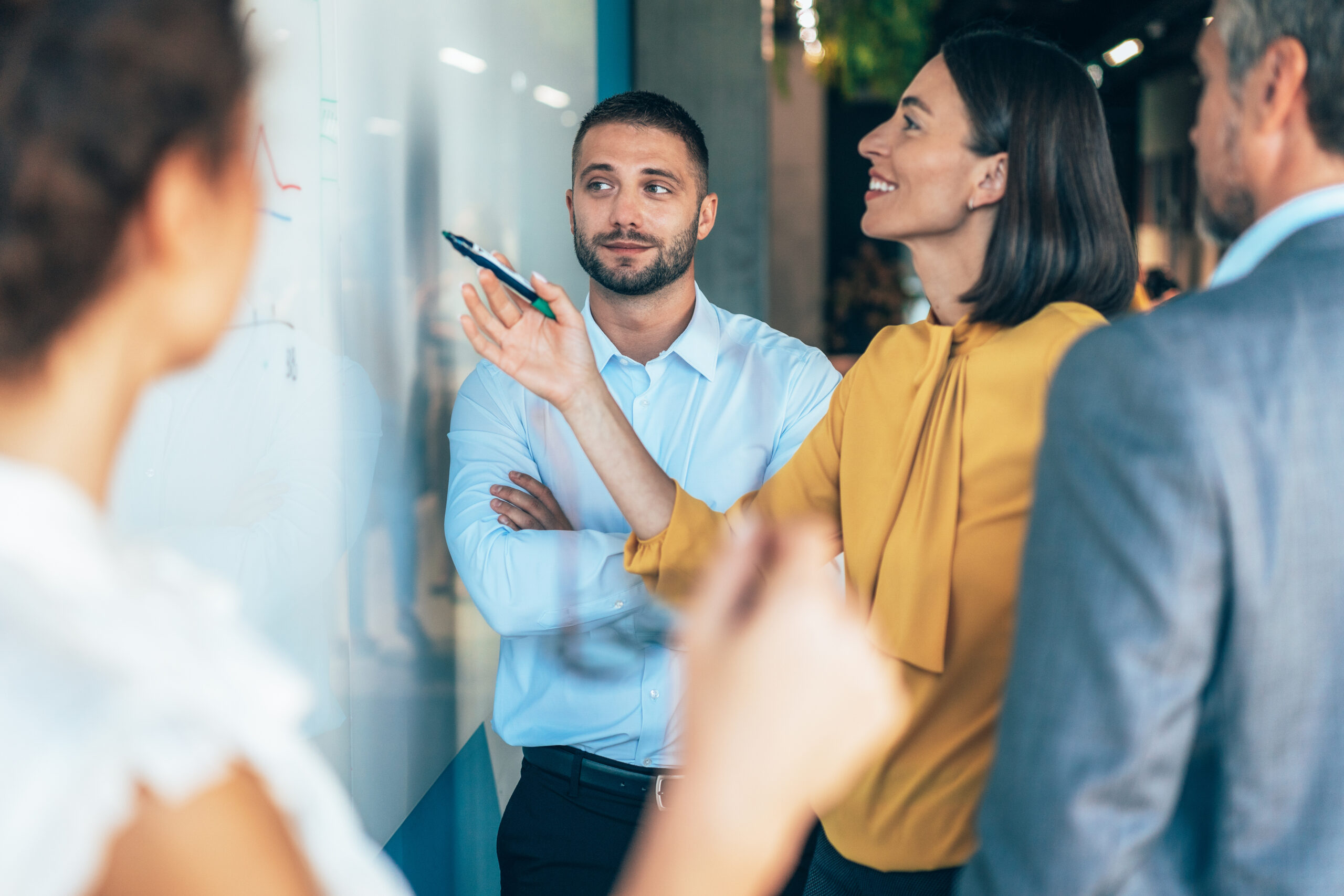 Colleagues in an animated discussion at a glass whiteboard, one holding a marker.
